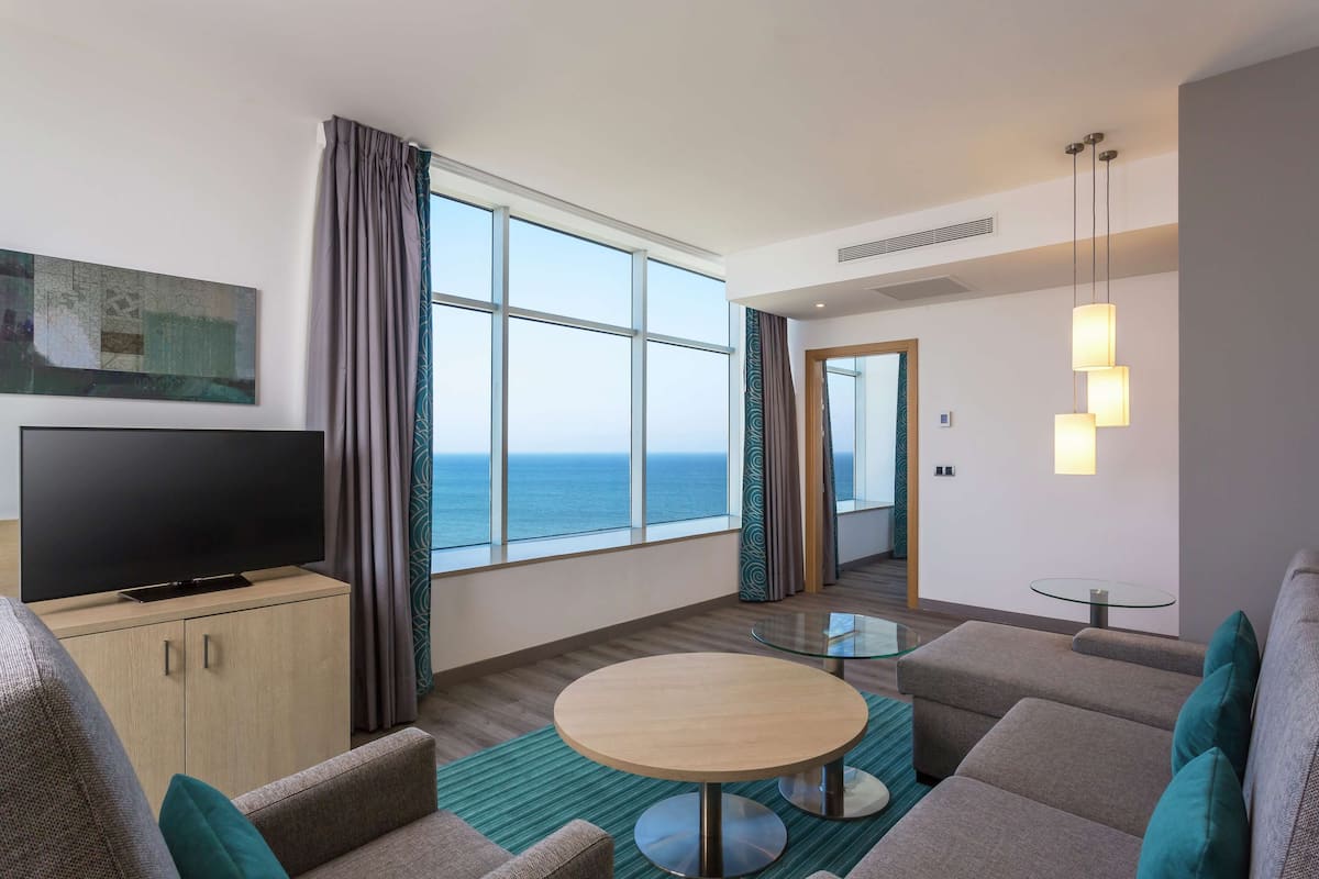 A modern hotel room with a large window offering a sea view, a flat-screen TV on a wooden cabinet, a round glass table, and a sofa with teal cushions.