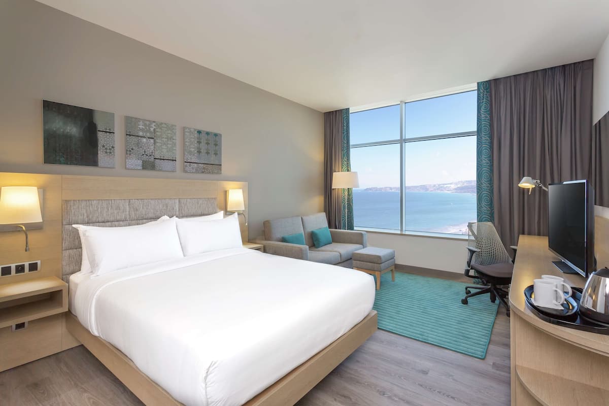 A hotel room with a large bed, a desk with a TV, a chair, and a sofa. There is a window with a view of the sea.