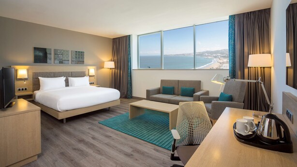 A modern hotel room with a large bed, a sofa, a desk with a kettle, and a view of the sea. - Hilton Garden Inn Tanger City Center (Tangier)