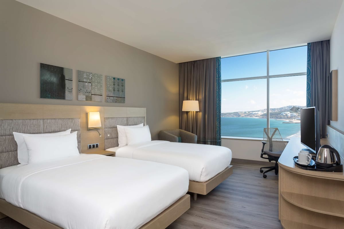 A hotel room with two beds, a desk with a computer, a chair, and a large window offering a view of the sea.