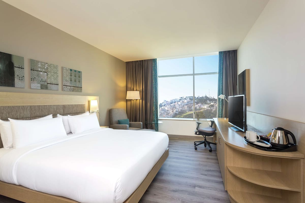 A hotel room with a large bed, a desk with a TV, and a view of a cityscape.