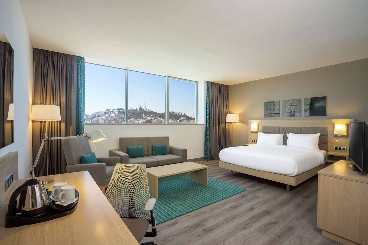 A hotel room with a bed, a sofa, a desk with a kettle and cups, a TV, and a city view.