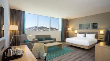 Habitación de hotel con cama, sofá, escritorio con tetera y tazas, televisor y vista a la ciudad.