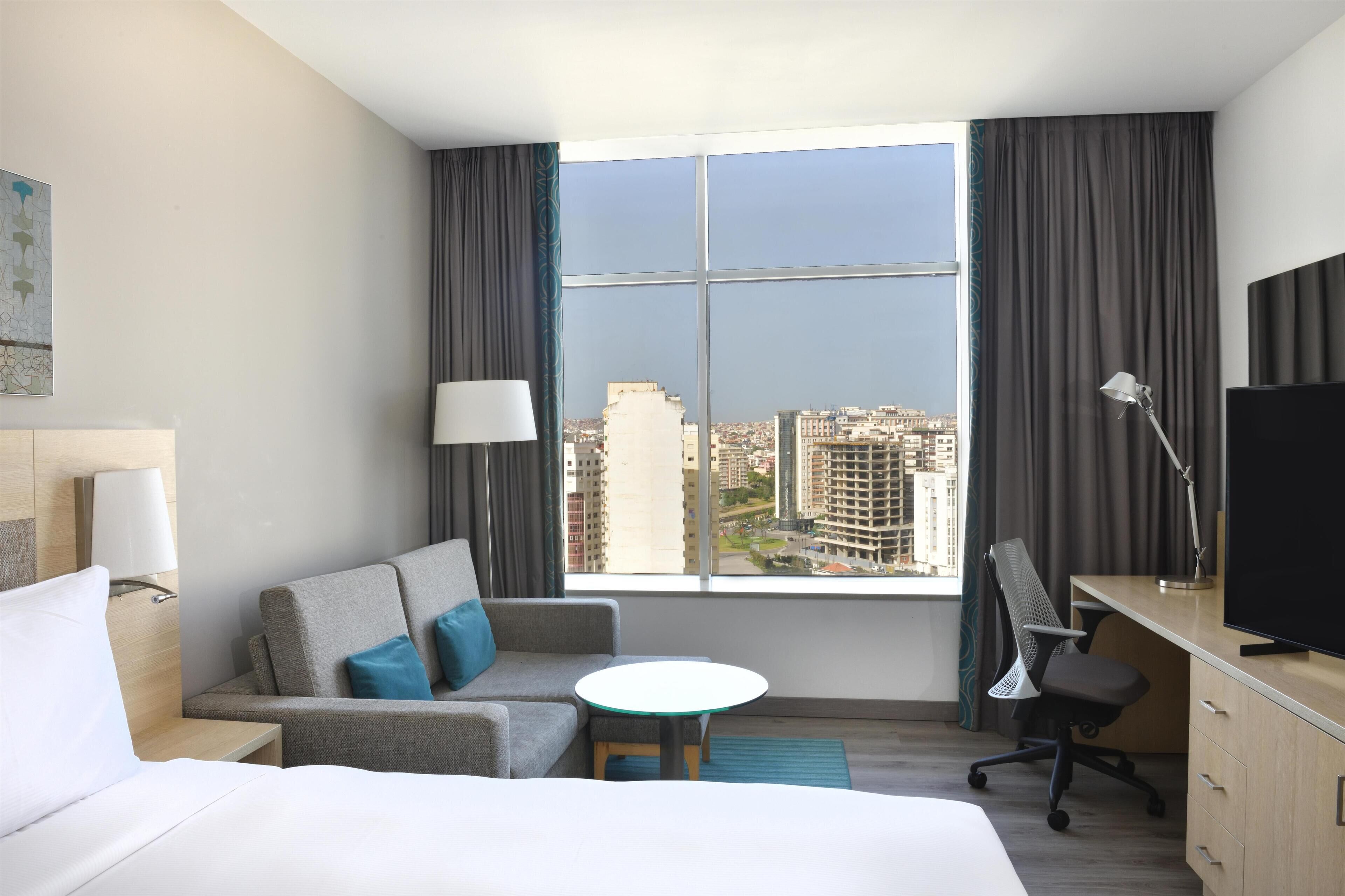 Habitación de hotel con cama, sofá, una mesita, escritorio, silla, televisor y una ventana grande con vistas a la ciudad.