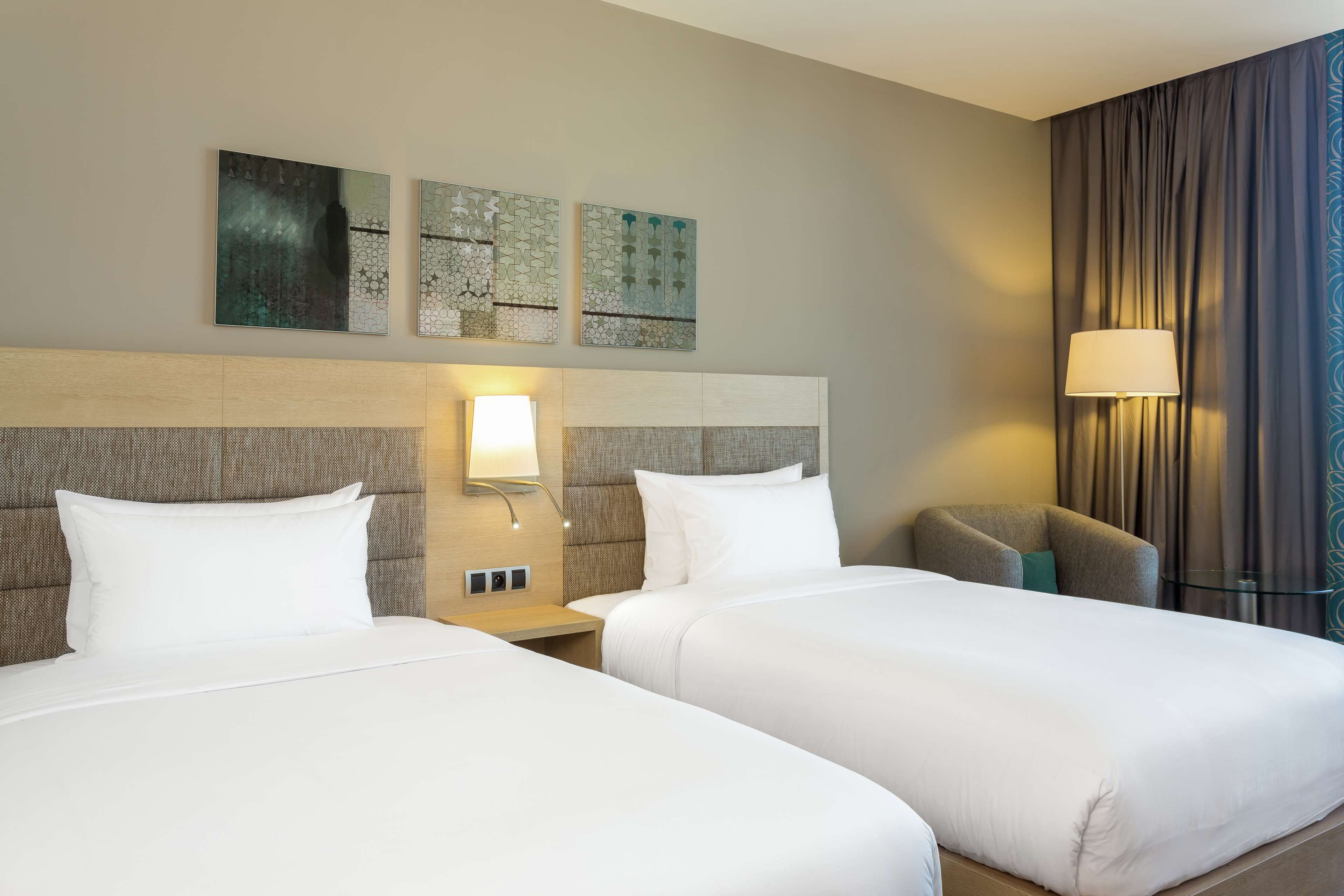 A hotel room with two beds, a headboard, a nightstand, a lamp, and a wall-mounted artwork.