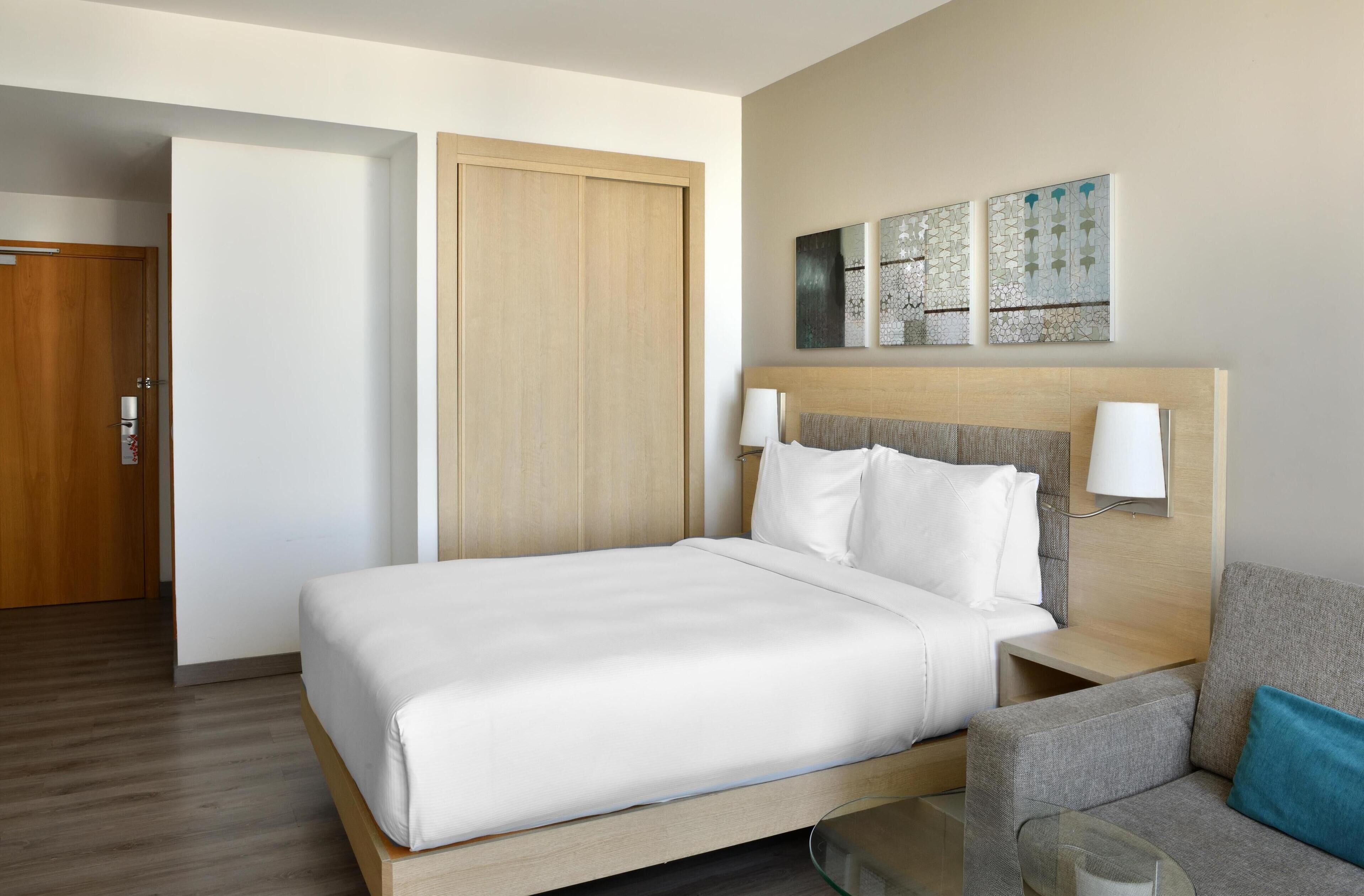 A hotel room with a bed, a nightstand, a lamp, a sofa, and artwork on the wall.