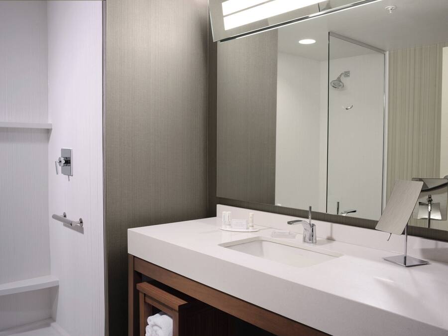 A modern bathroom with a white sink, a large mirror, and a shower visible in the reflection.