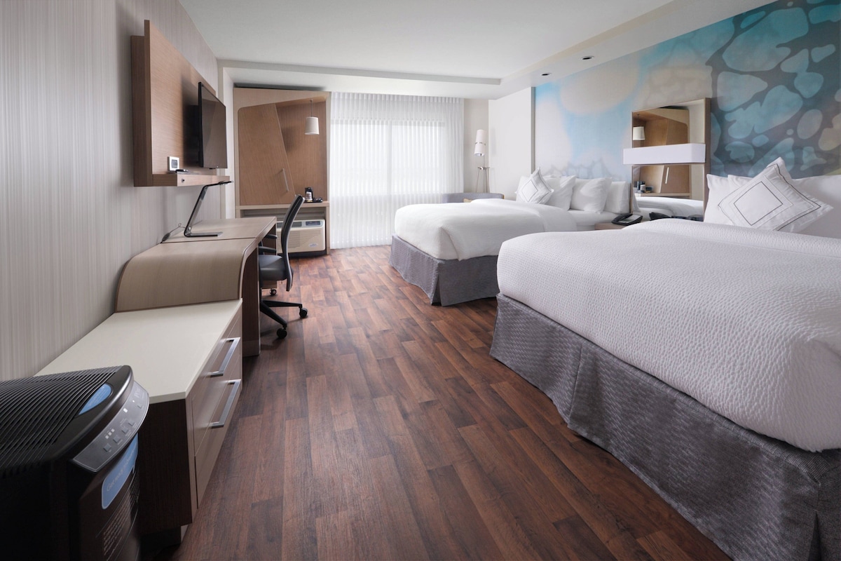 A hotel room with a wooden floor, a desk with a chair, a bed with a grey bed skirt, a TV mounted on the wall, and a painting on the wall.