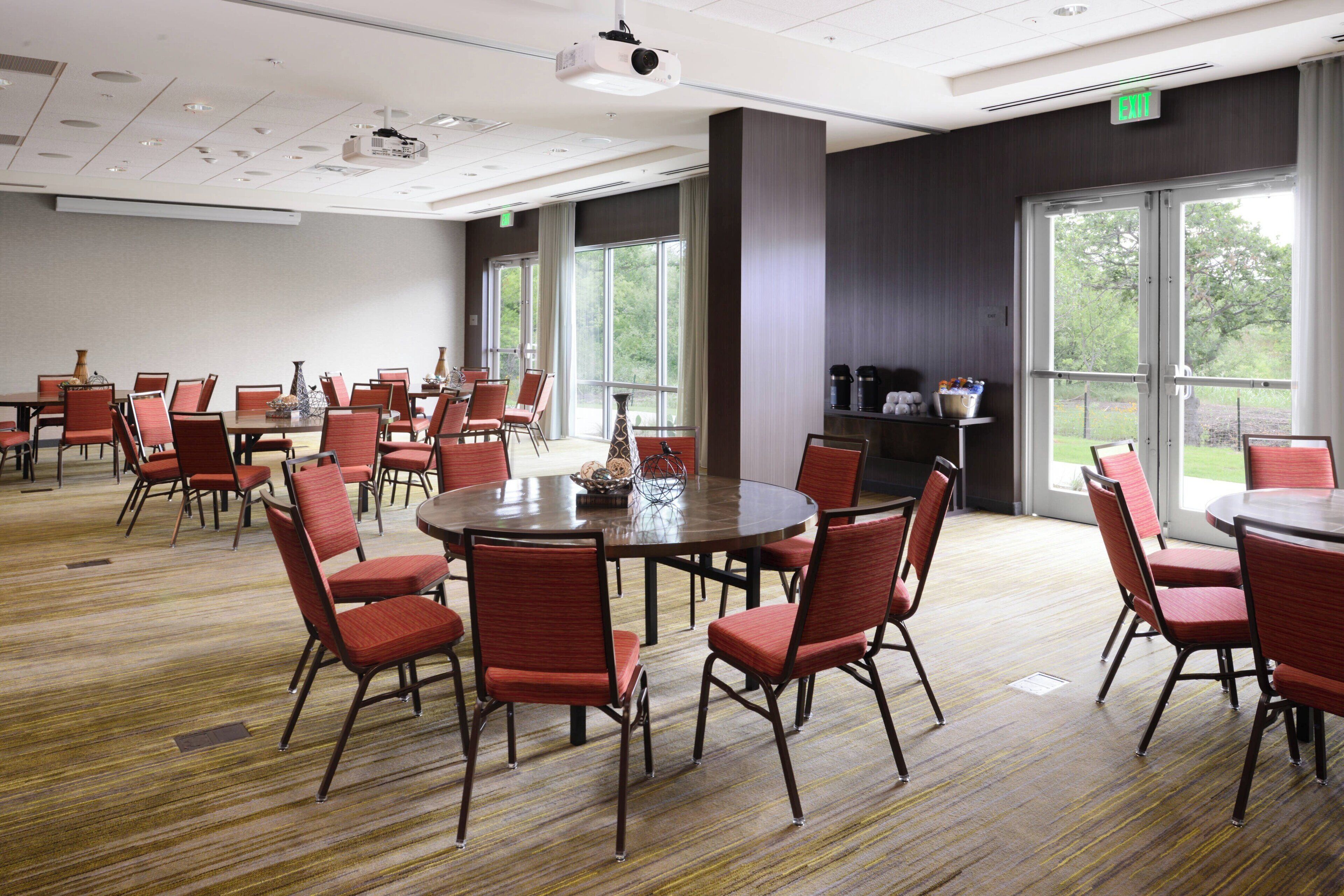 A conference room with multiple tables and chairs, large windows, and a projector on the ceiling.