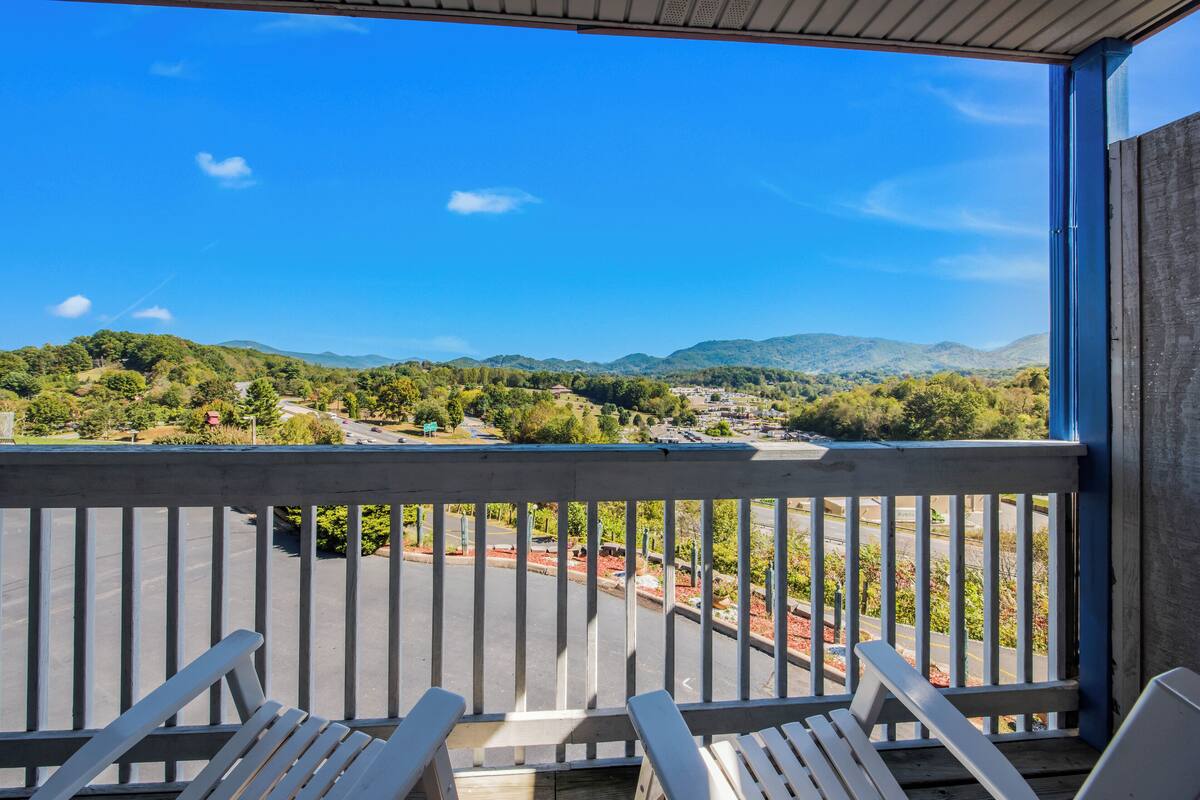 A balcony with white railings and two white chairs, overlooking a road and green hills.