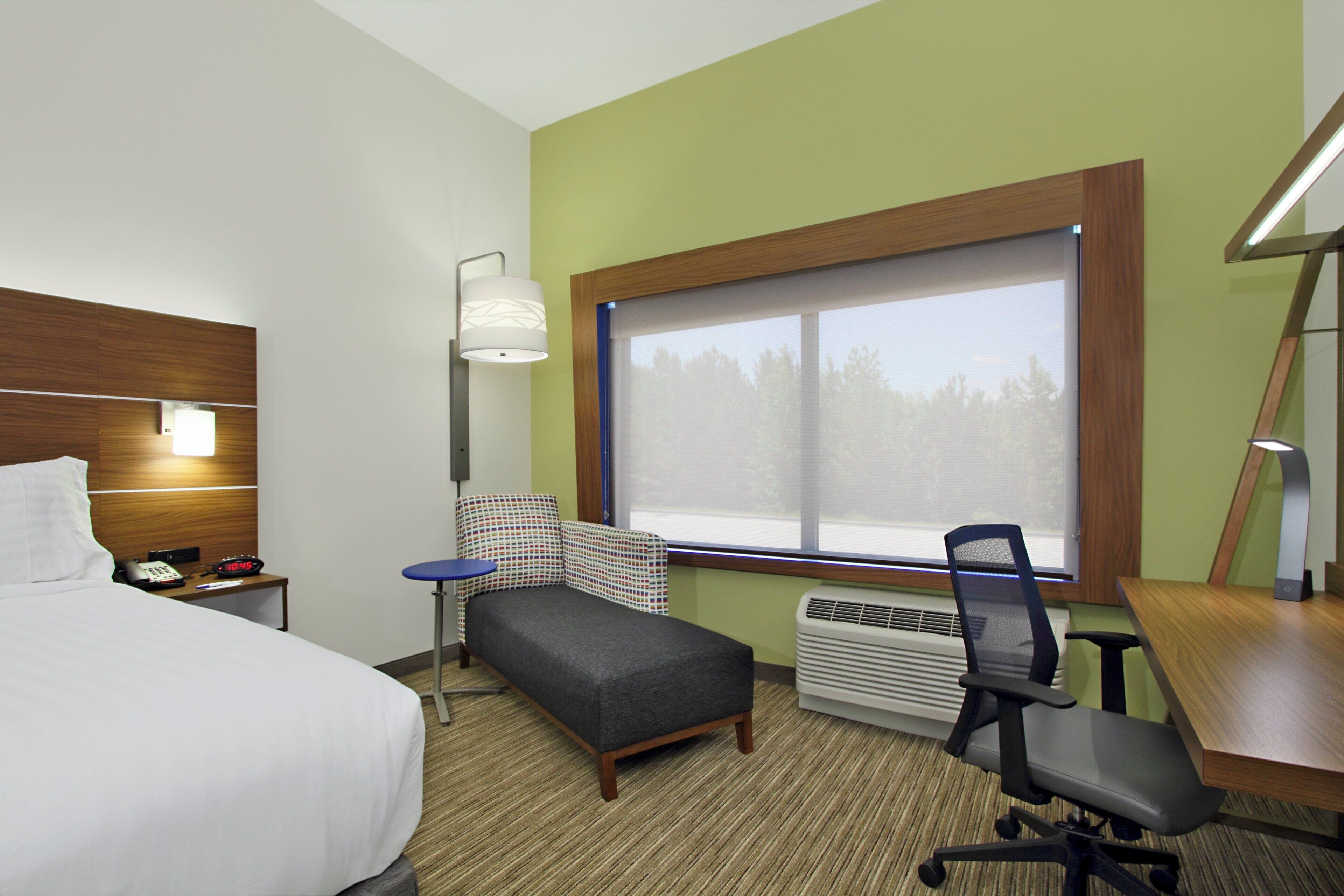 Habitación de hotel con cama, escritorio, silla, una mesita, lámpara, calefactor y ventana con vista a árboles.