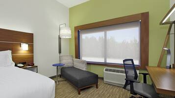 Habitación de hotel con cama, escritorio, silla, una mesita, lámpara, calefactor y ventana con vista a árboles.