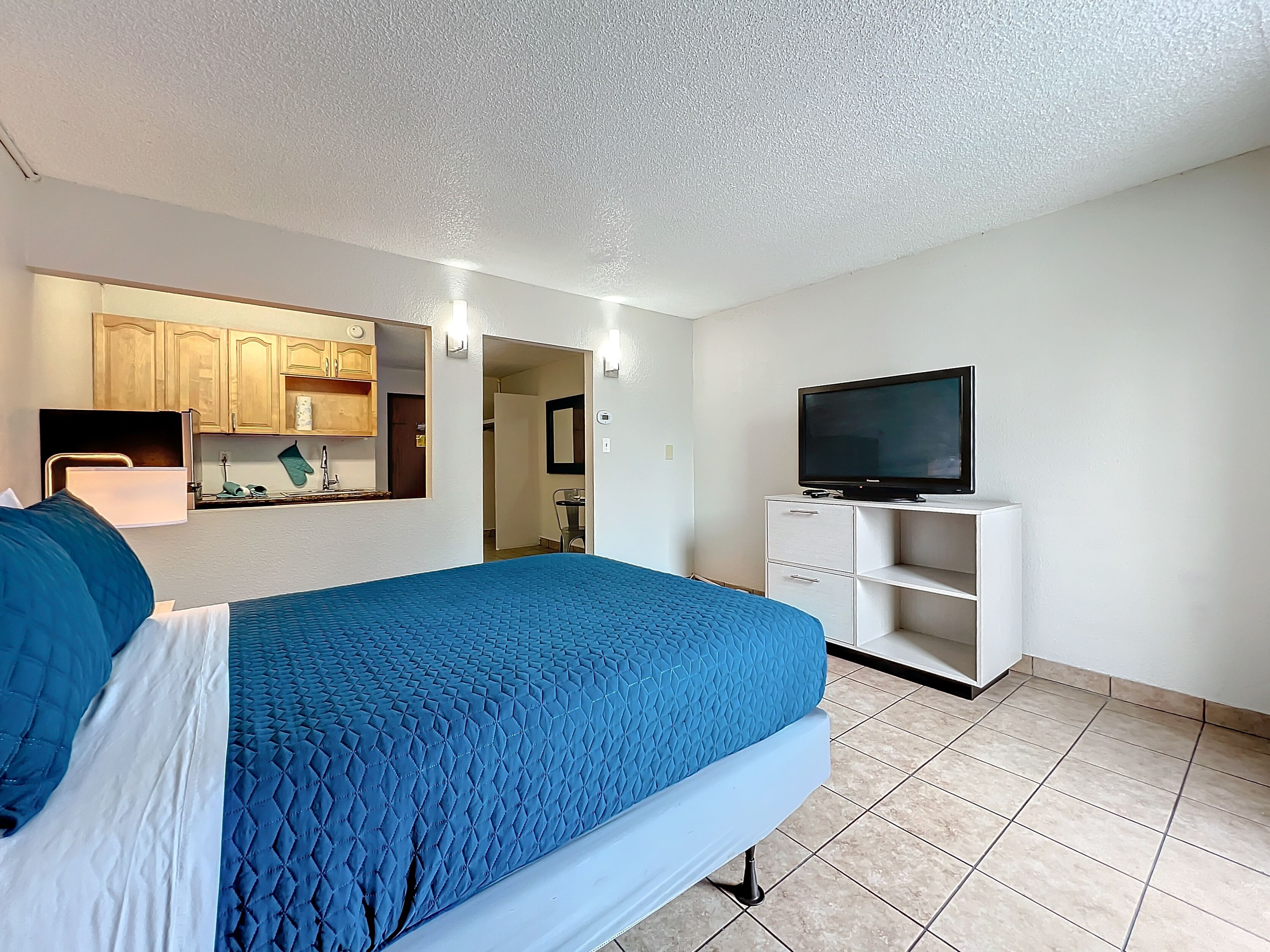 A hotel room with a bed, a TV on a stand, a kitchenette with cabinets, and a bathroom visible through an open door.