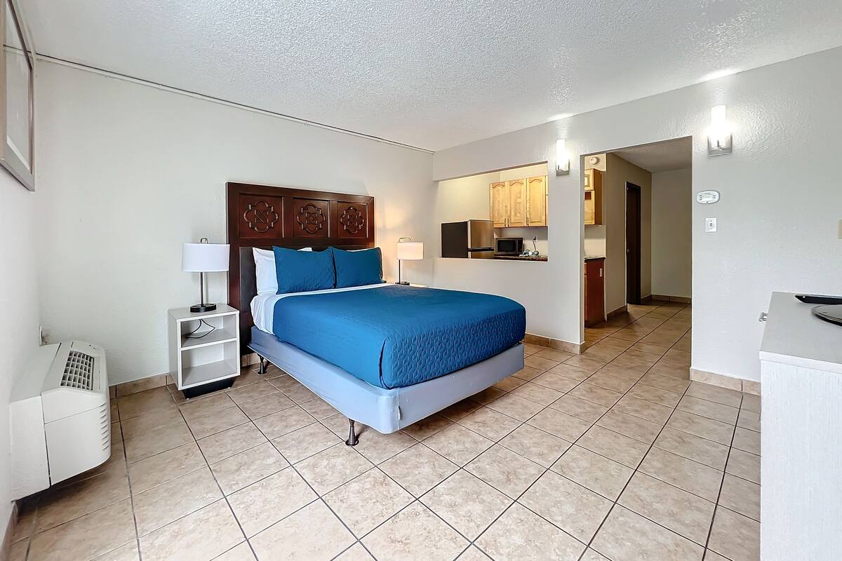 A hotel room with a bed, a nightstand, a lamp, a small white cabinet, and a kitchenette in the background.