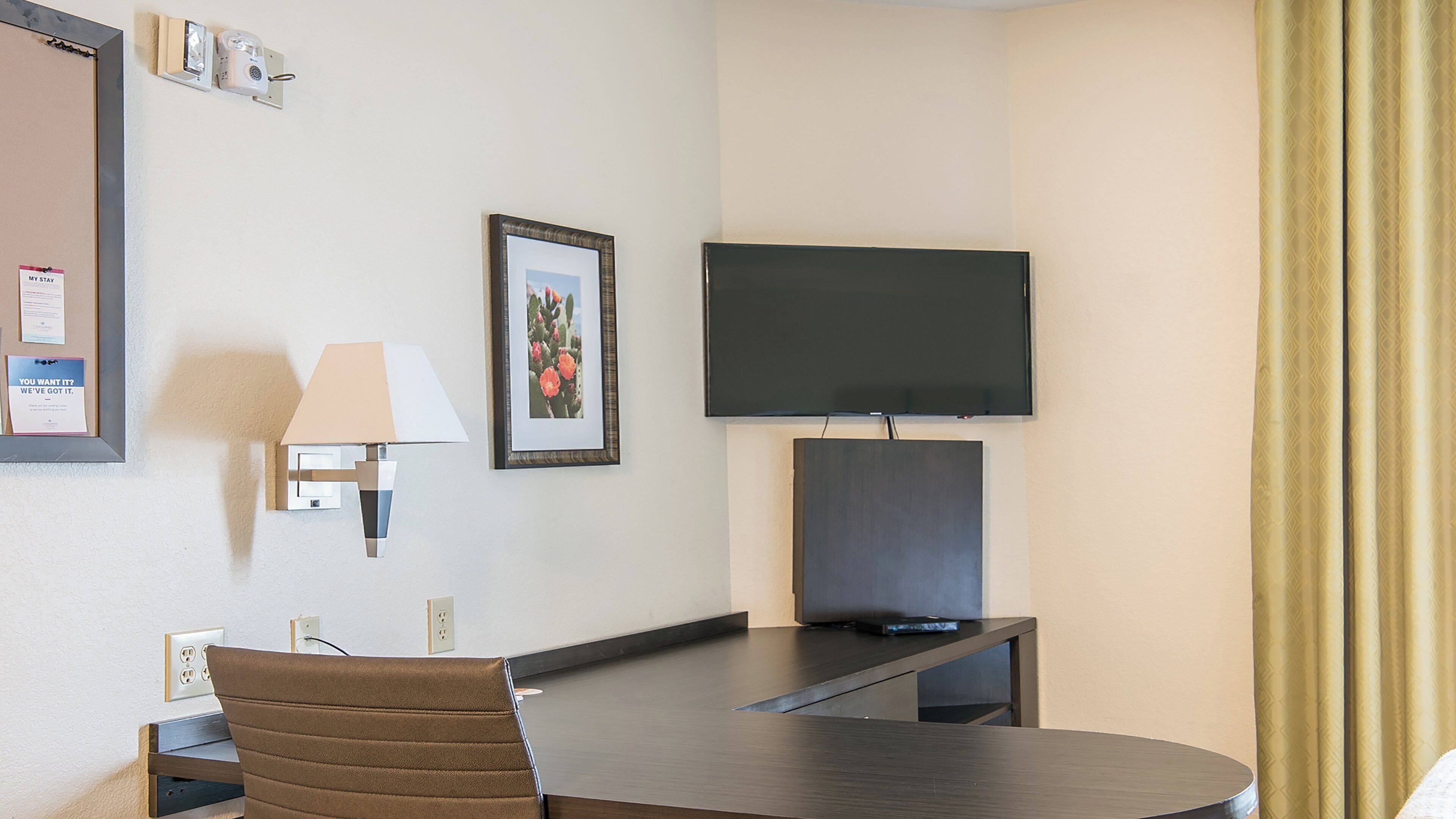 A hotel room with a bed, a desk with a chair, a television mounted on the wall, and a framed picture.