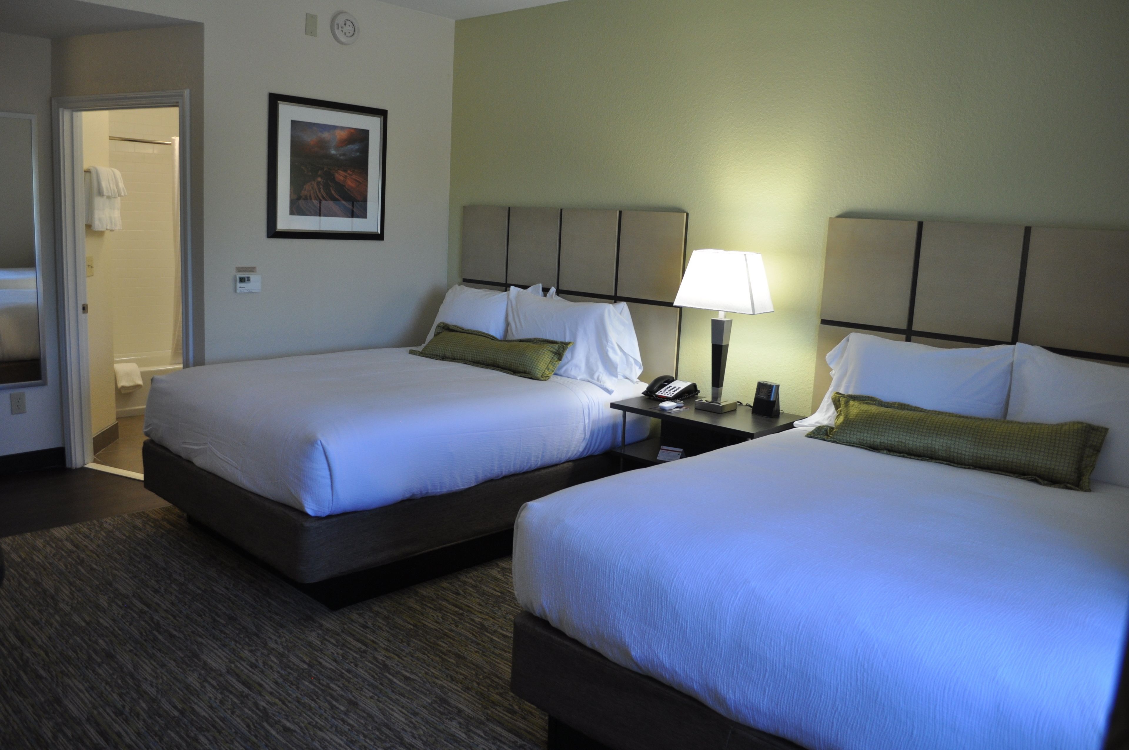 A hotel room with two beds, a nightstand with a lamp, a framed picture on the wall, and a door leading to a bathroom.
