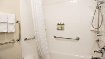 Une salle de bain avec des murs carrelés de carreaux blancs, une baignoire, une douche avec un pommeau de douche à main et des barres d’appui. Deux bouteilles de shampoing et d’après-shampoing sont posées sur le mur.
