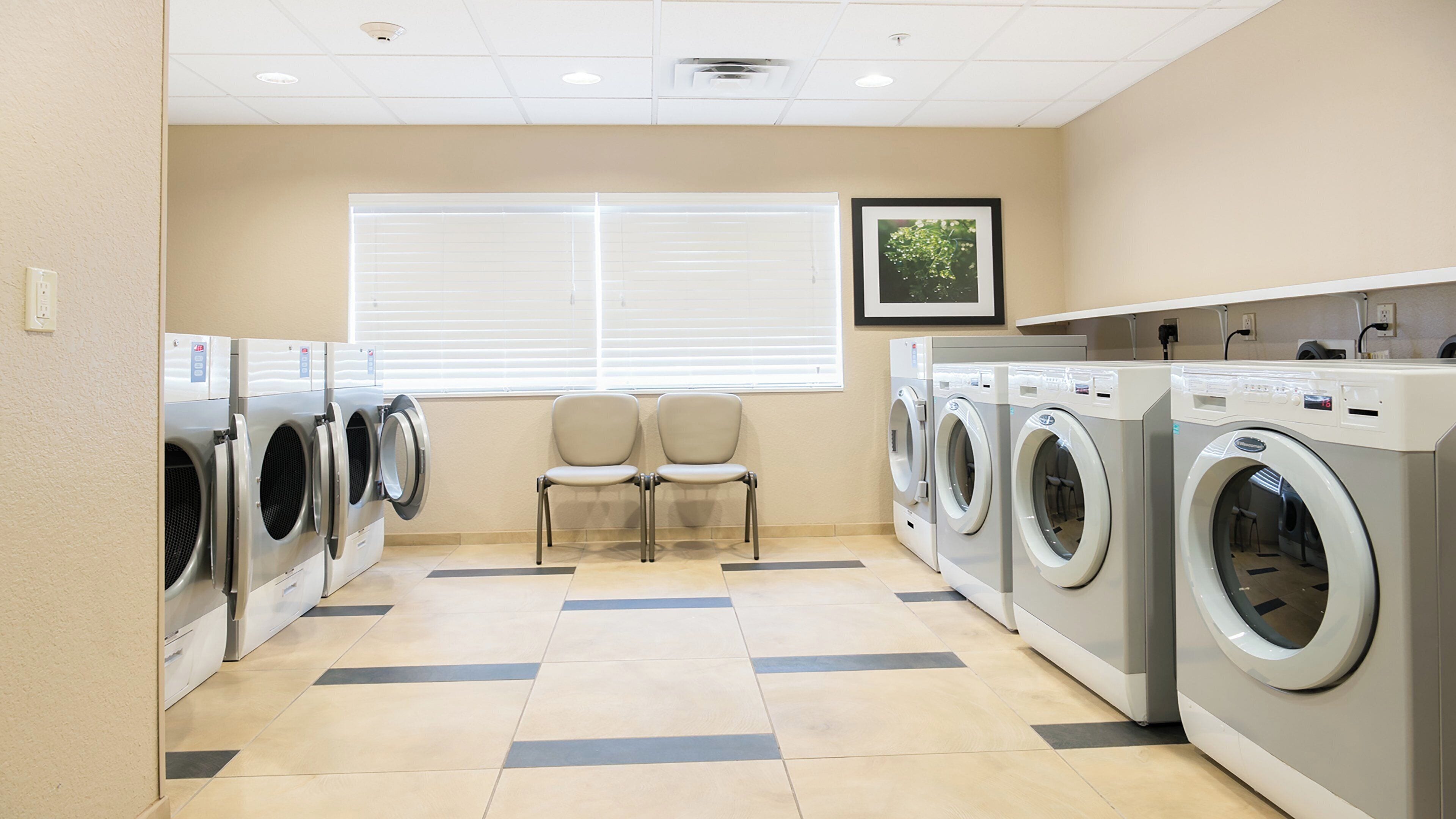 A laundry facility with multiple washing machines, chairs, and a large window with blinds.