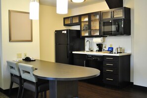 Cocina compacta con refrigerador, microondas y cafetera. Pequeño comedor con dos sillas y una mesa.