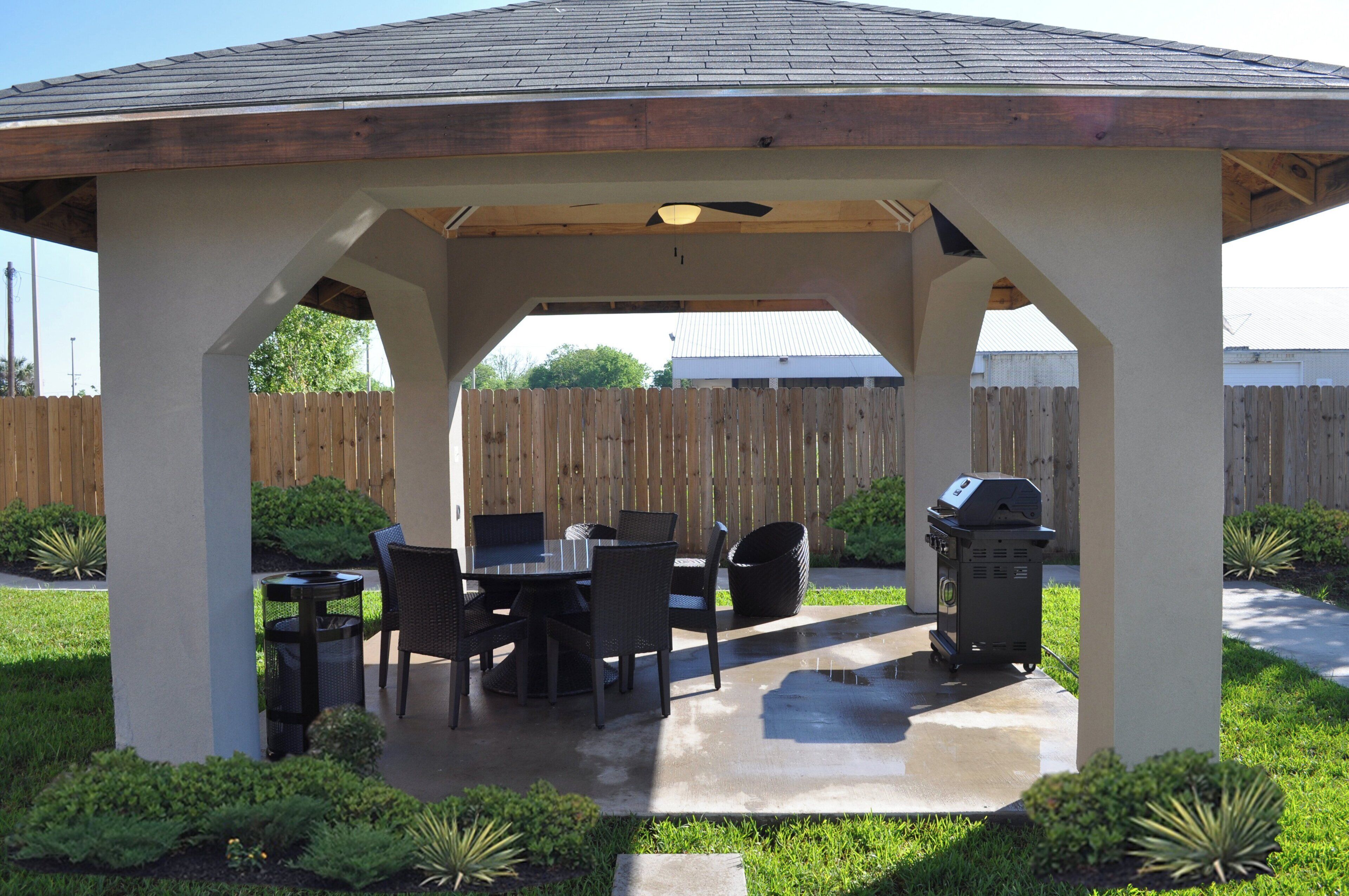 A covered patio with a table and chairs, a grill, and a wooden fence in the background.