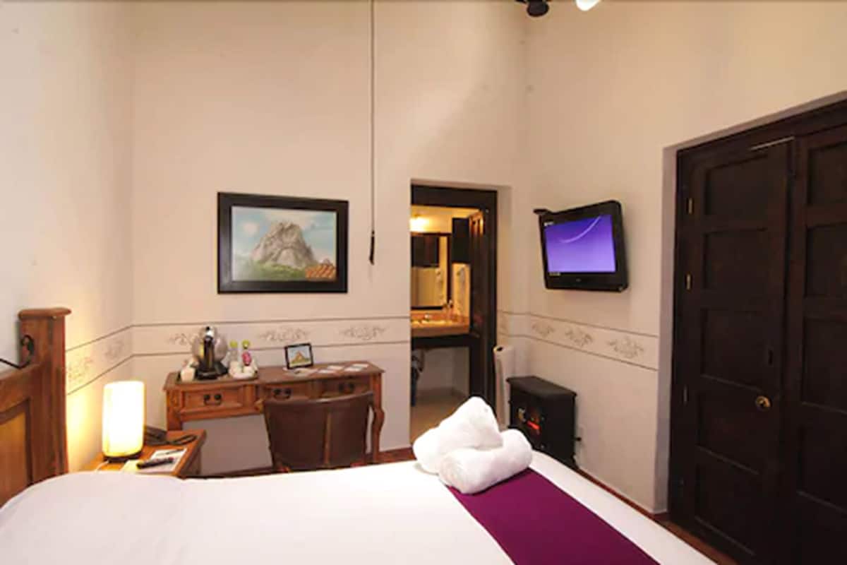 A hotel room with a bed, desk, chair, lamp, TV, and a door.