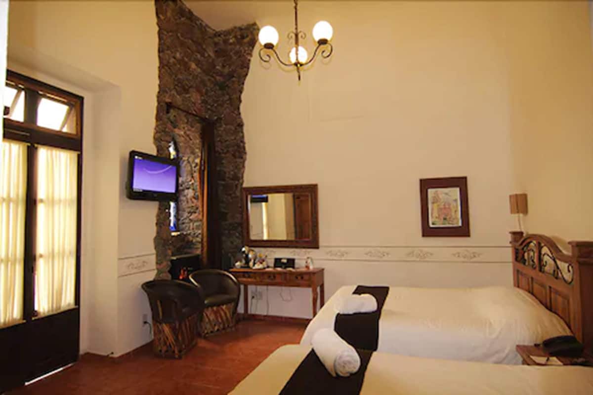 A hotel room with a large stone wall, a television, a desk, a bed, and a chandelier.