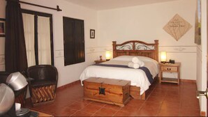 A bedroom with a wooden bed, a nightstand with a lamp, a chest of drawers, a chair, and a window with curtains. - Hotel Villa del Villar (Querétaro)