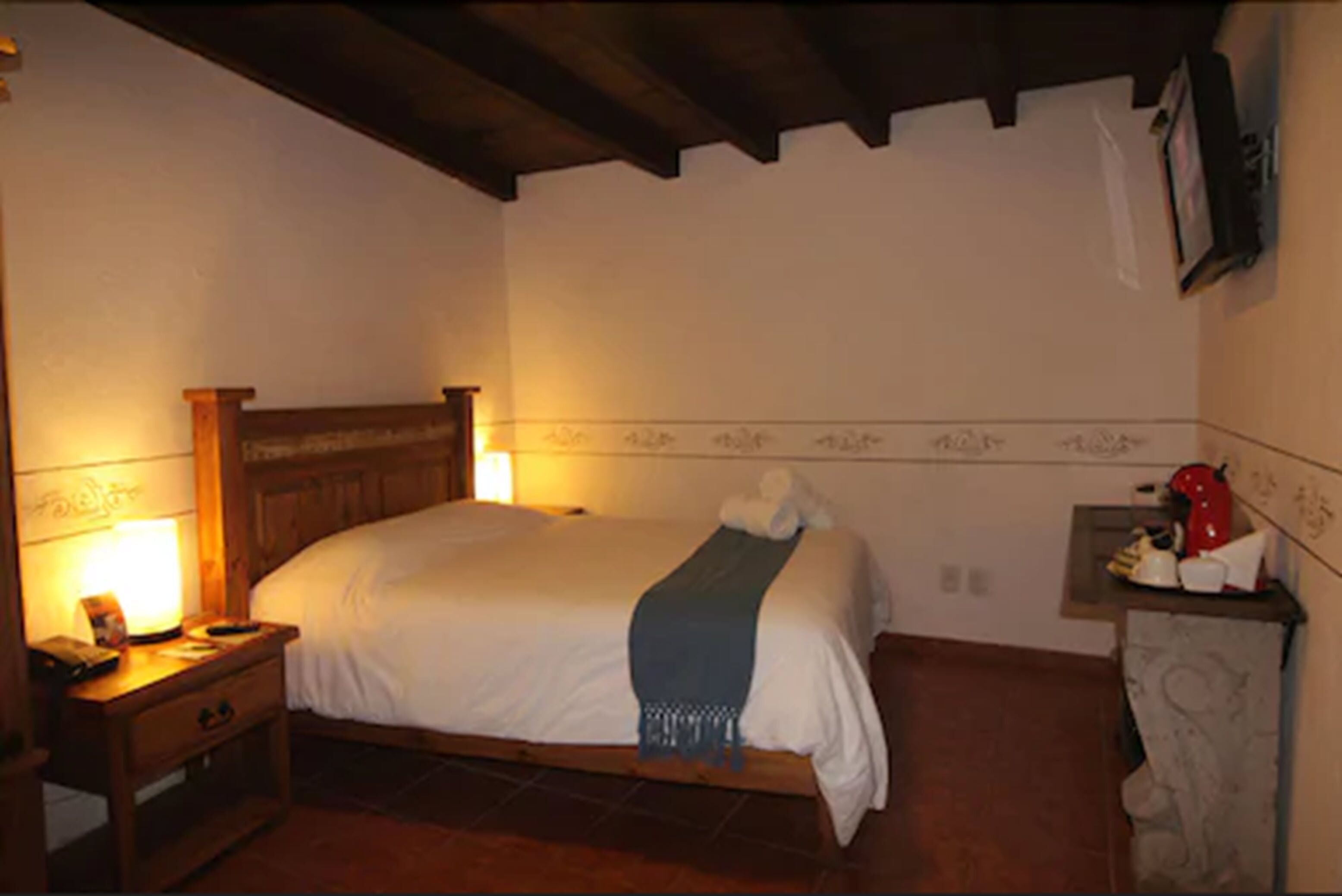 Un dormitorio con una cama de madera, una mesita de noche con una lámpara, una pequeña mesa con un objeto rojo y un televisor montado en la pared.