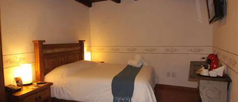 Un dormitorio con una cama de madera, una mesita de noche con una lámpara, una pequeña mesa con un objeto rojo y un televisor montado en la pared.