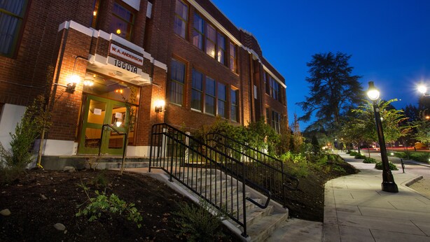 A brick building with a sign reading "W.A. Anderson 18607A" at the entrance, a staircase with a metal railing, and a well-lit sidewalk. - McMenamins Anderson School (Bothell)