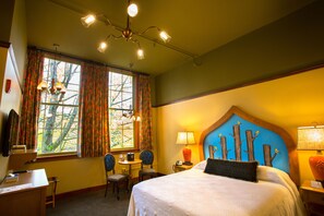 A hotel room with a large bed, a desk with a chair, a small table, and a window with curtains. - McMenamins Anderson School (Bothell)