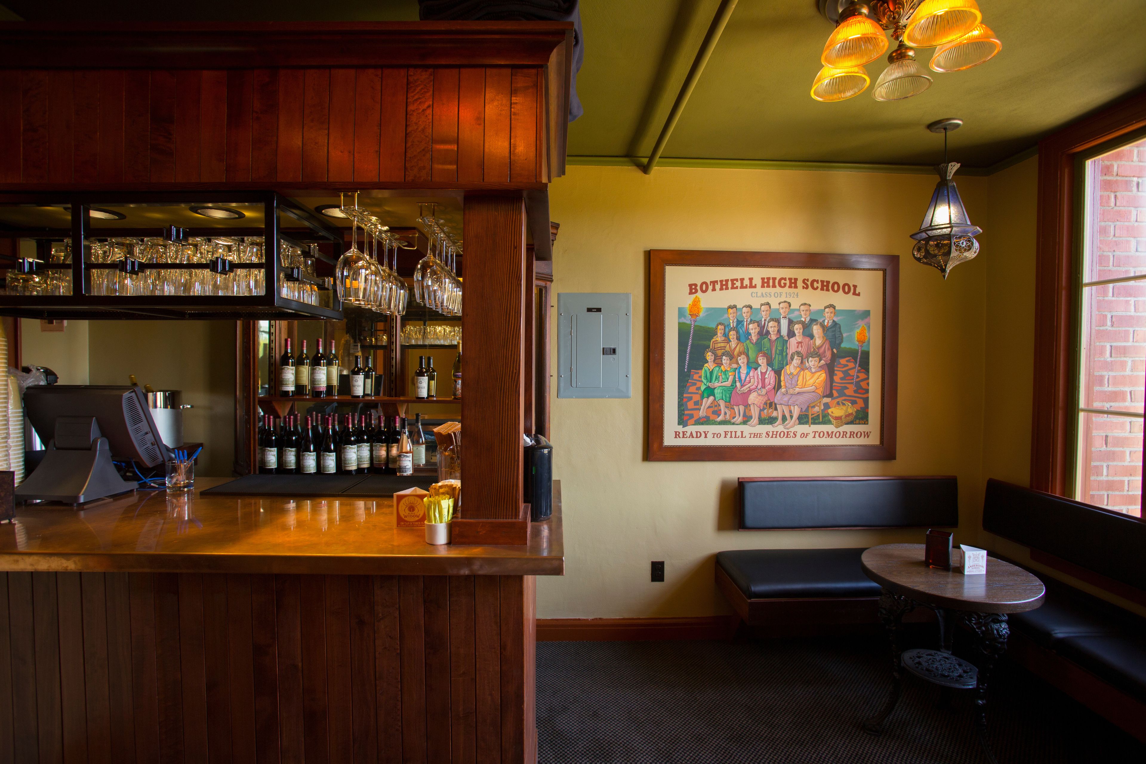 Una zona de bar con estantes de madera, botellas y vasos. Hay un cartel enmarcado en la pared y un banco con una mesa.