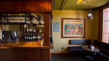 Eine Bar mit Holzböden, Flaschen und Gläsern. An der Wand hängt ein gerahmter Plakat, und es gibt eine Bank mit einem Tisch.