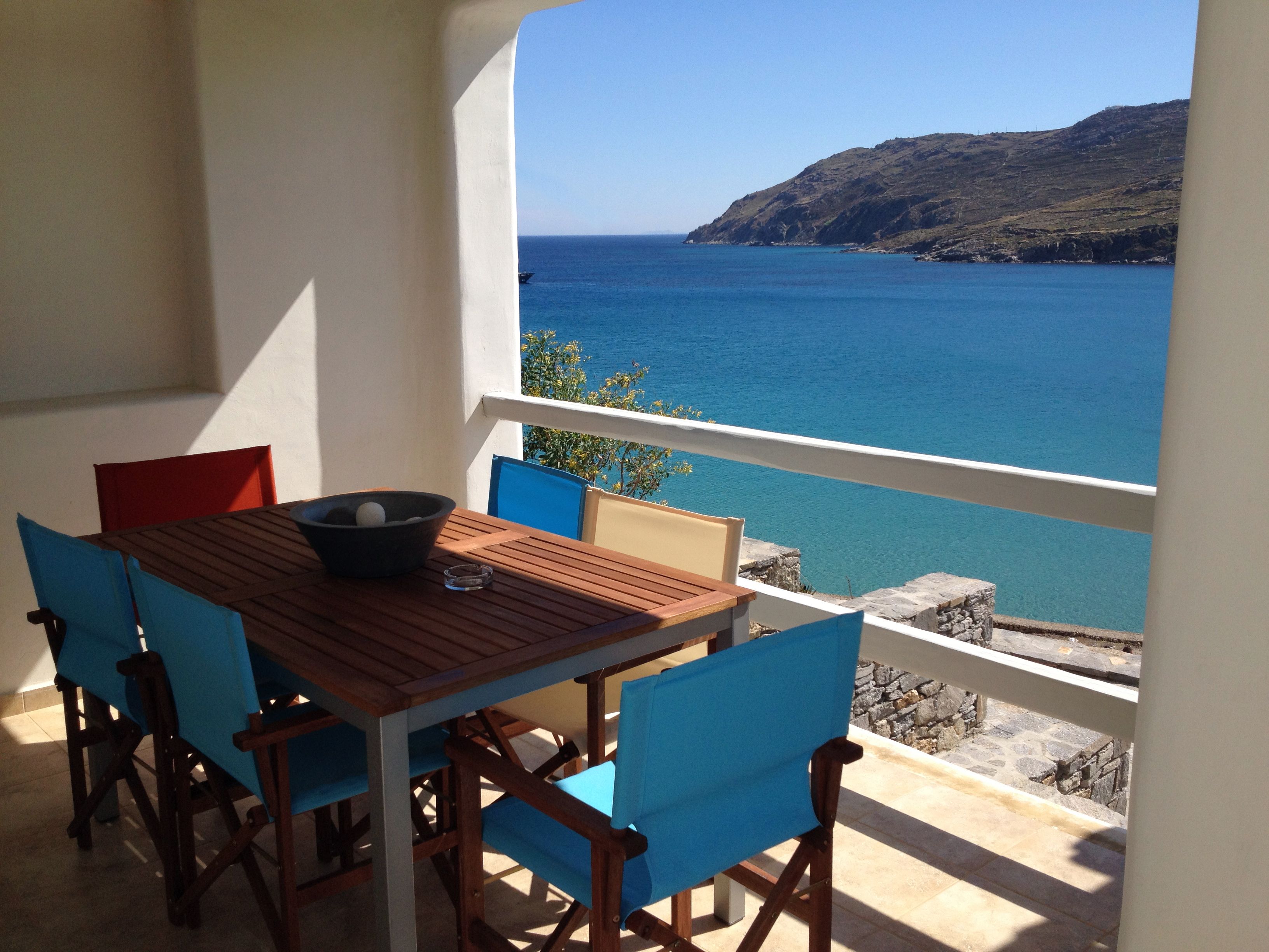 A wooden table with blue chairs on a balcony overlooking a clear blue sea and a hilly landscape.
