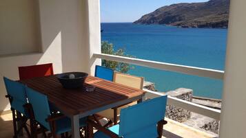 A wooden table with blue chairs on a balcony overlooking a clear blue sea and a hilly landscape.
