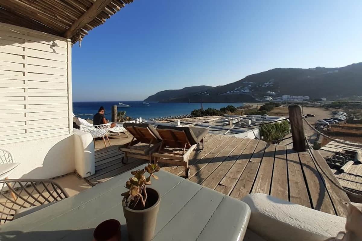A wooden deck with lounge chairs and a table, overlooking a coastal view with hills and buildings in the distance.