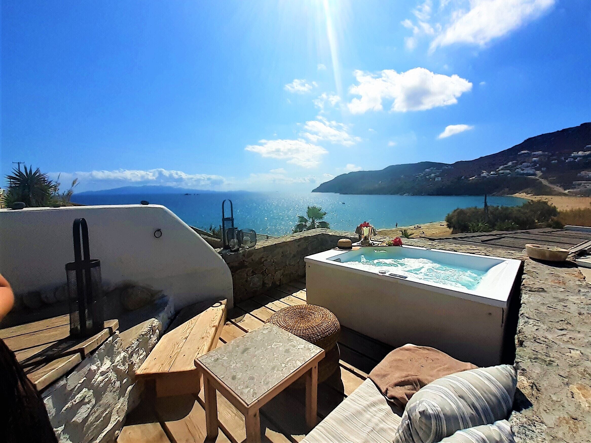 A terrace with a hot tub, wooden furniture, and a view of the sea and mountains.