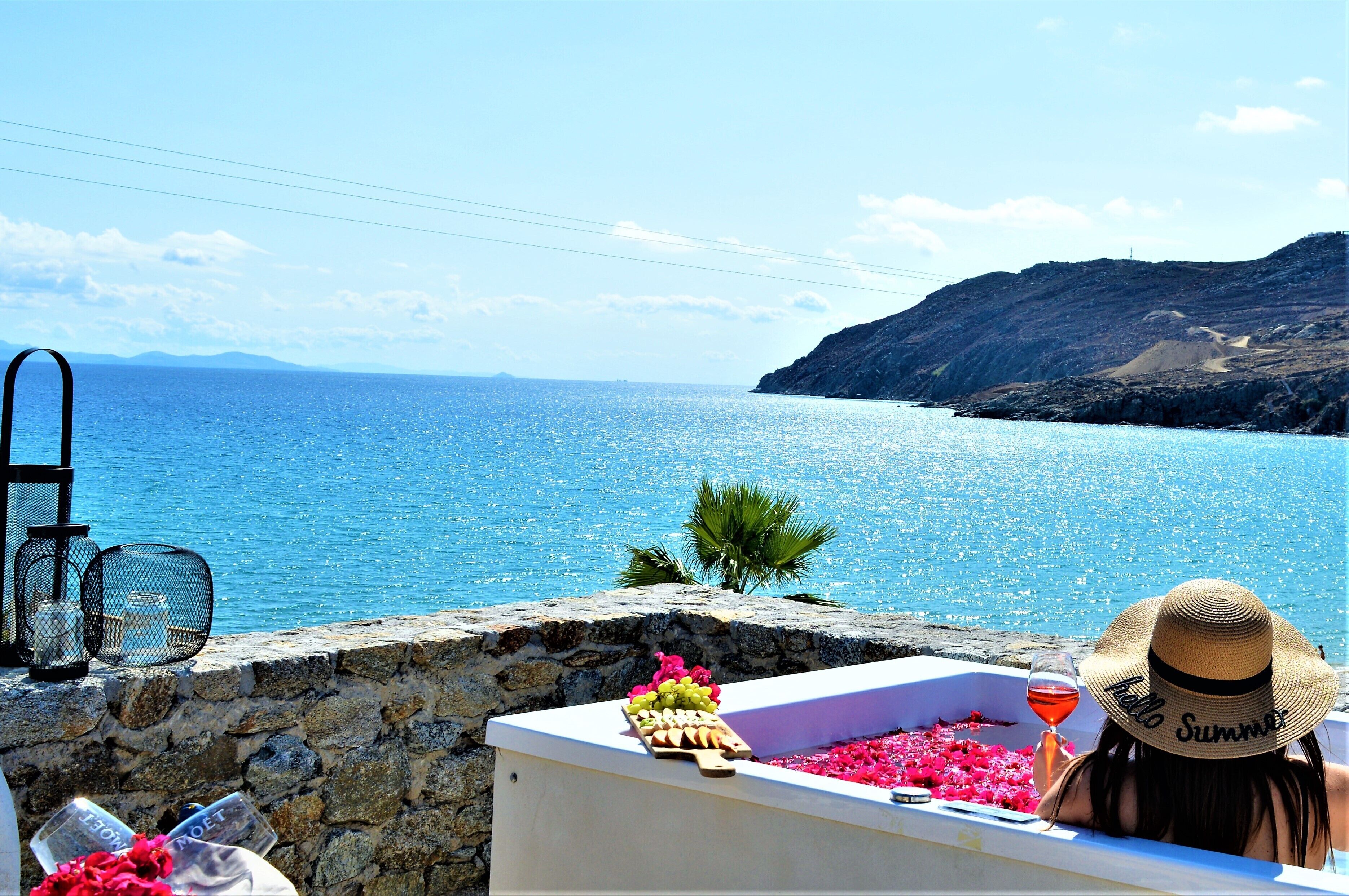 Vista costera con un muro de piedra, una bañera blanca con flores y una copa de vino, y un sombrero de paja con el texto 'Hello Summer'.