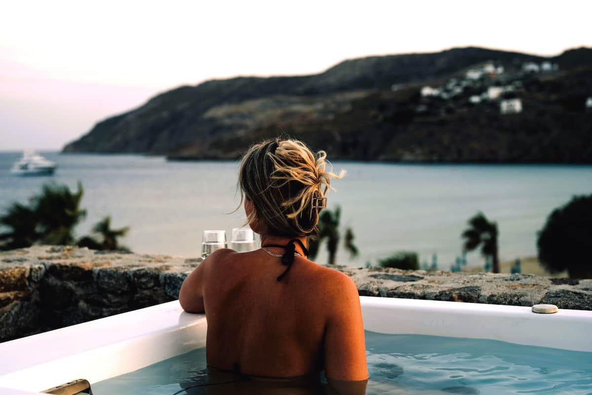 A person in a bathtub overlooking a body of water with a boat in the distance and a mountainous landscape.