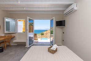 Un dormitorio con cama, televisión, ventana con vista al mar y una cesta de mimbre con una toalla.