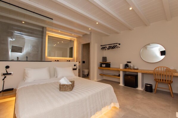 A modern bedroom with a large bed, a bedside table, a mirror, a wooden chair, and a small desk with a computer and a coffee machine. - Aelia Mykonos (Mykonos)
