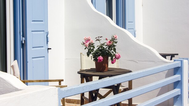 A white building with blue doors and shutters, a round wooden table with a potted plant, and a blue railing. - Sourmeli Garden (Mykonos)