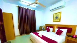 Habitación de hotel con una cama, ventilador de techo, aire acondicionado, un cuadro en la pared y una mesita de noche de madera.