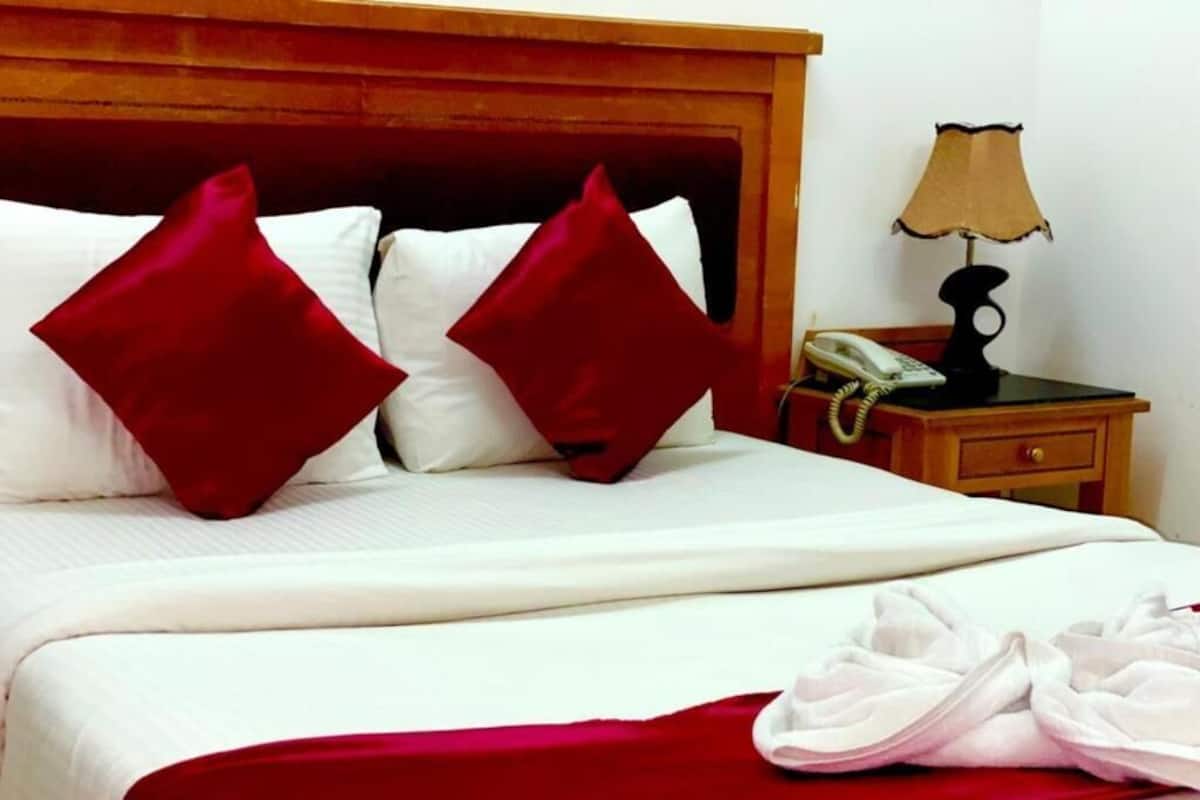 A neatly made bed with red pillows, a white blanket, and a red throw. There is a bedside table with a lamp and a telephone.