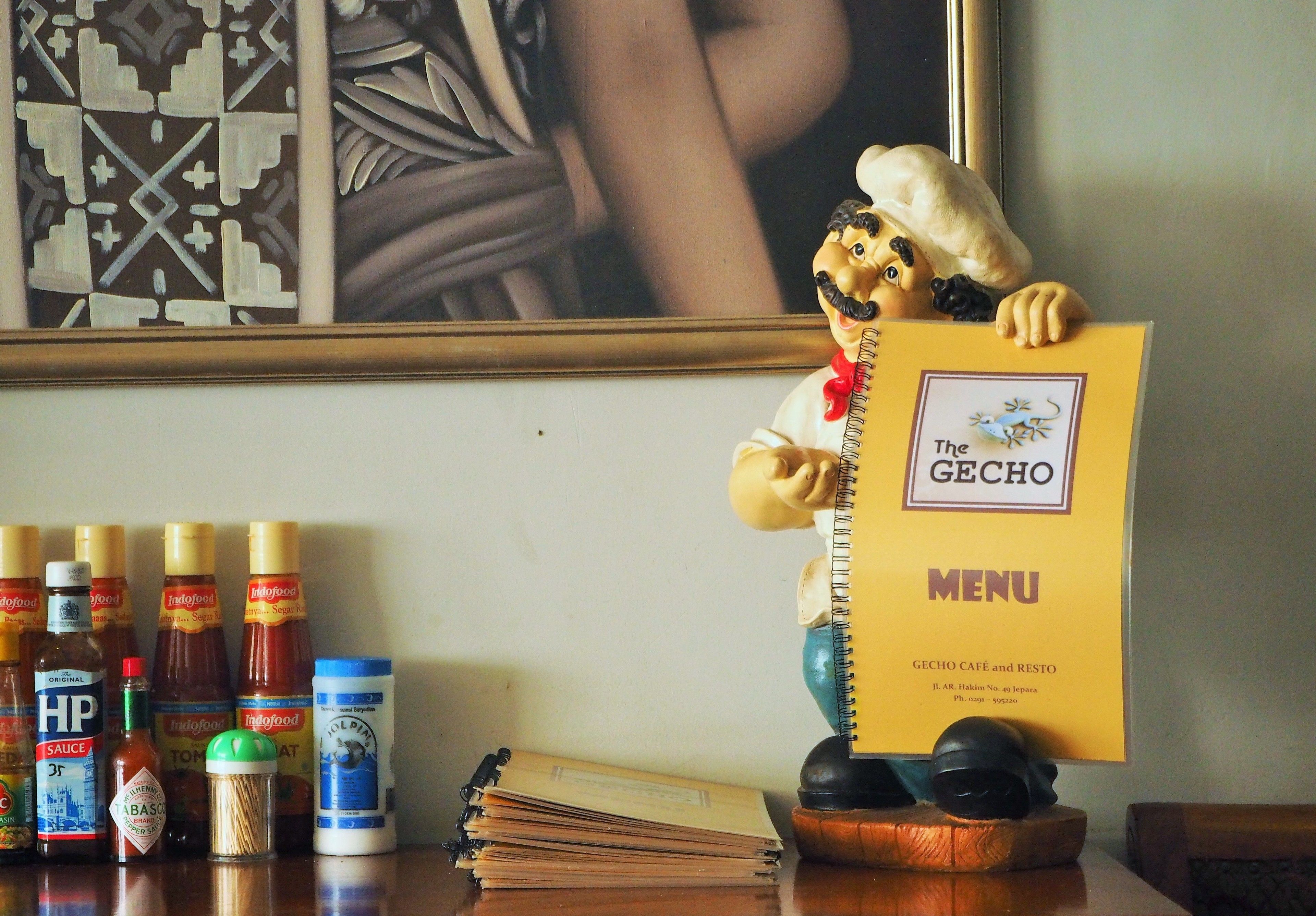 Une statuette représentant un chef tenant un menu, entouré de condiments et d’une pile de livres posés sur une table.