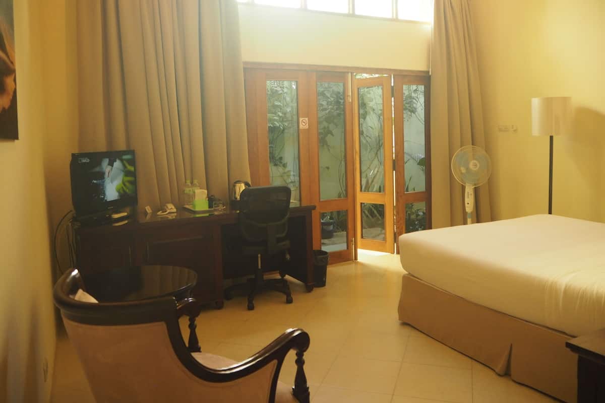Habitación de hotel con cama, escritorio con computadora, silla, bote de basura y ventana con cortinas.