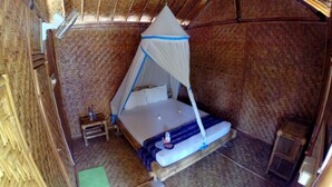 A single bed with a mosquito net in a room with bamboo walls and a thatched roof. - Same Same - Bungalows - Hostel (Kuta)