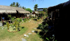 A tropical garden with thatched-roof structures, stone pathways, and various plants. - Same Same - Bungalows - Hostel (Kuta)