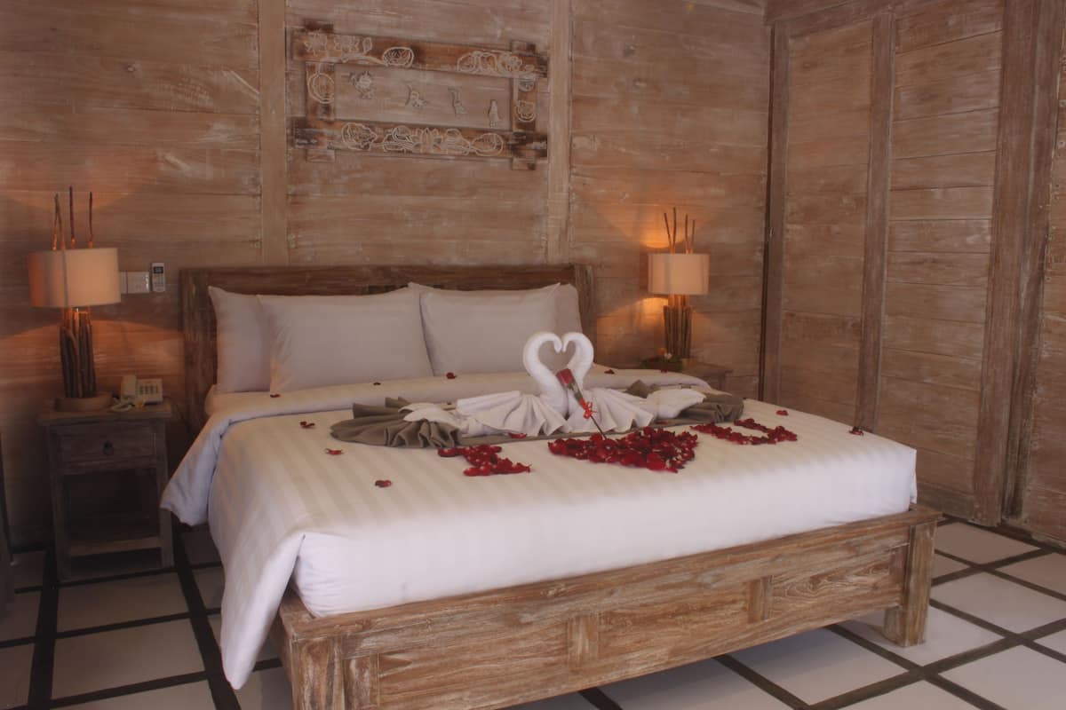 A neatly made bed with a heart-shaped towel arrangement, two bedside lamps, and a wooden headboard.