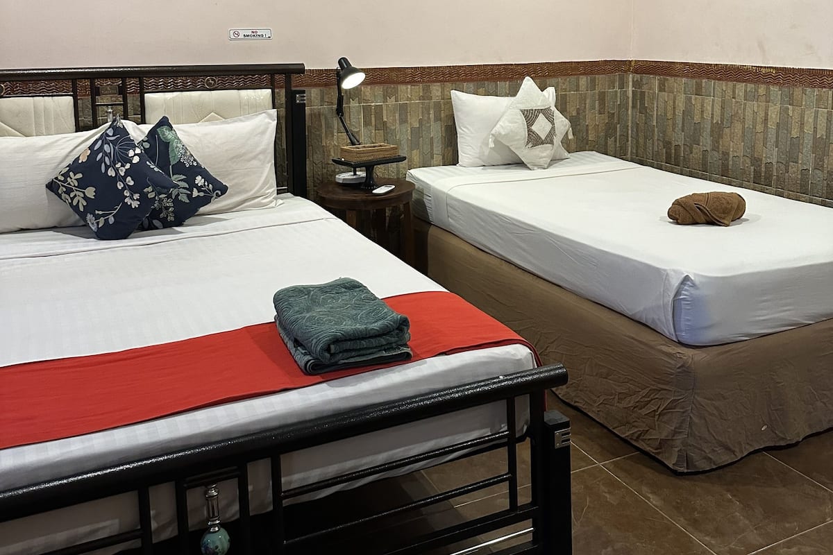 A hotel room with two single beds, a nightstand, a lamp, a framed picture on the wall, and a 'No Smoking' sign.